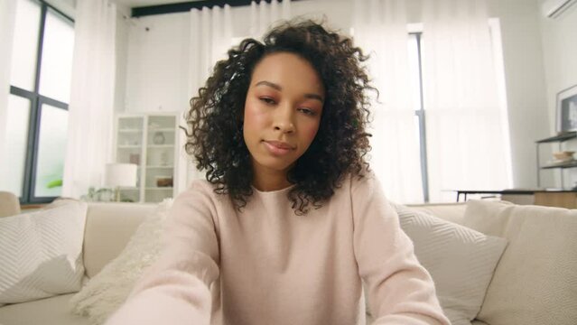Puzzled young African American woman making selfie or video call while sitting at home on sofa. Portrait of concerned, uncertained and frustrated female with curly hair doing video chat in apartment