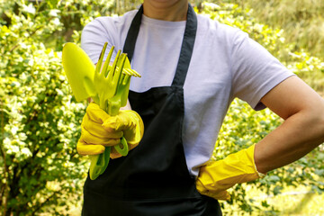 a woman in an apron and household gloves holds gardening tools in her hands on a summer day