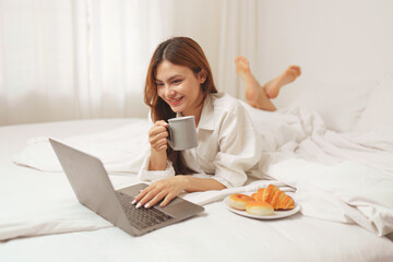 Happy smiling asian woman stretching on bed in the morning and using laptop computer