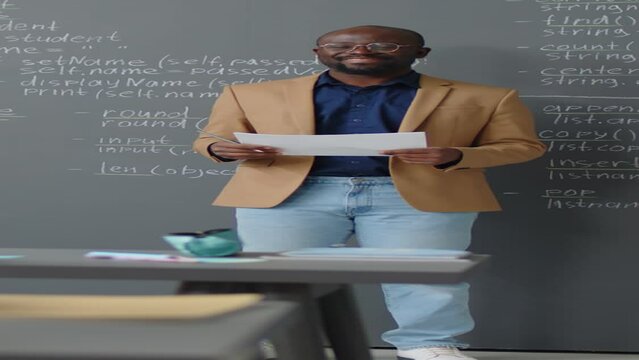 Vertical full shot of male African American teacher standing in front of blackboard with code written on it giving lecture