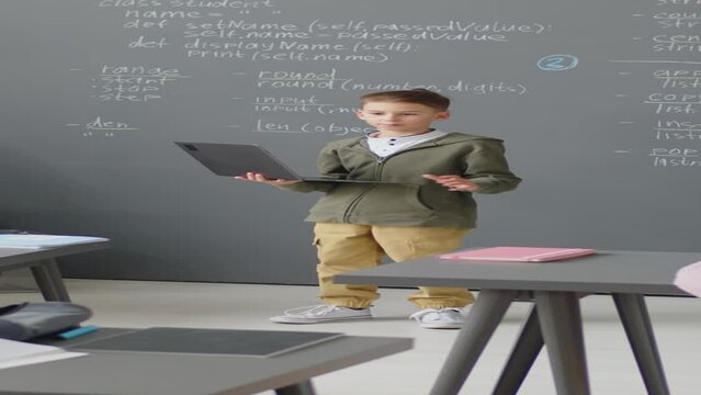 Full shot of elementary age Caucasian boy standing in front of class holding laptop and making report at daytime