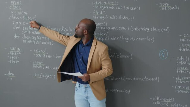 Medium shot of male African American teacher standing in front of blackboard in classroom explaining code description at daytime