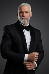 Attractive middle-aged man with grey hair and beard is dressed in a stylish black suit and bowtie, portraying a suave secret agent during a photoshoot against a grey backdrop - obrazy, fototapety, plakaty
