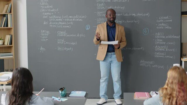 Full shot of male African American teacher standing in front of board in classroom explaining new material to ethnically diverse female students at daytime