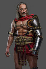 Aged but still mighty, this muscular gladiator with long grey hair and beard stands proudly in lightweight armor against a grey background