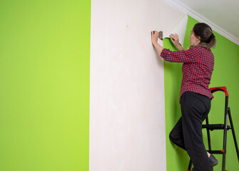 A woman on a stepladder near a wall with torn wallpaper. Wallpaper change, renovation in the apartment and at home. A woman performs wall repairs