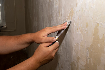 A spatula in men's hands removes old plaster from the wall in the house. The concept of cleaning...