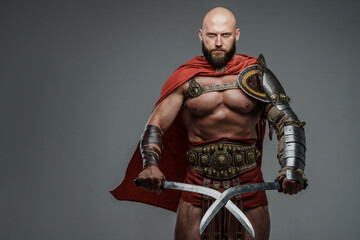 Bald and bearded gladiator poses standing tall with two swords while wearing light armor. This fearsome and imposing warrior emanates power and strength against a neutral gray background