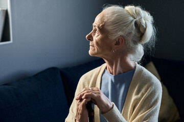Lonely senior woman with grey hair put in bun looking through window while sitting in front of...