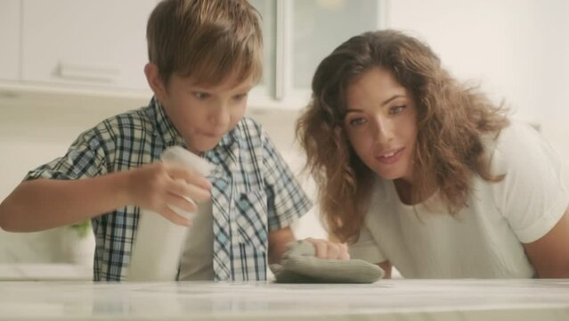 Medium shot of pre-teen boy wiping table with rag and detergen under mothers instructions at kitchen