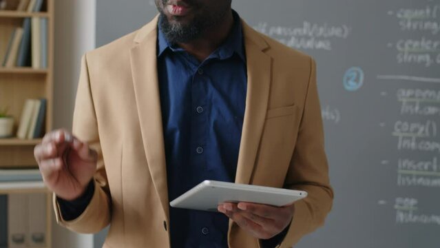 Tilt shot with focus on tablet in hands of male African American teacher walking there and back in front of students giving lecture on programming in school at daytime