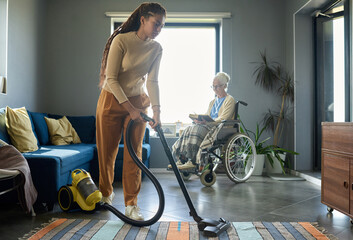 Cute teenage girl in casualwear cleaning floor of living room with vacuum cleaner against her...