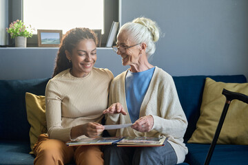 Happy old woman in eyeglasses looking at her granddaughter while sitting on sofa next to her and showing her album with family photos