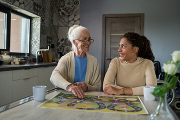 Cheerful teenage girl looking at smiling grandmother while sitting by table in the kitchen in front...