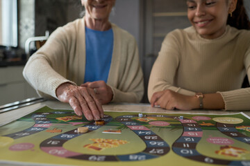 Hand of senior woman moving small round chip along board game while sitting by table next to her granddaughter and both playing at leisure