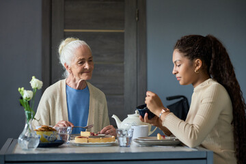 Kind grandmother looking at her granddaughter eating homemade food prepared by granny while sitting...