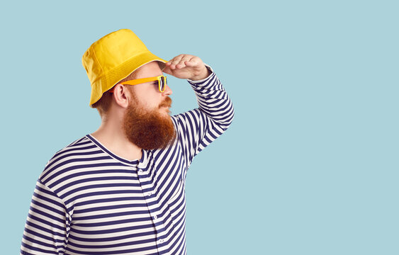 Funny fat man keeping hand on his forehead and with serious expression looking into distance on pastel light blue background. Profile view of man in summer yellow panama, sunglasses and sailor's shirt