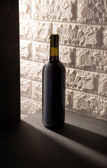 Unlabeled red wine bottle mockup on gray stone surface, white rustic brick wall background. Diagonal long shadows. Minimalist concept.