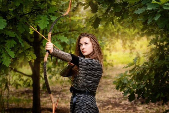 Medieval warrior woman in chain mail and bracers with a bow in her hands against the backdrop of an evening summer forest.