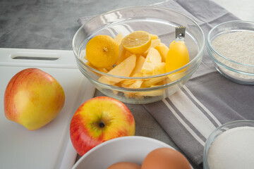Baking pan with apple slices, lemon, eggs, sugar, and flour in bowls close-up on a kitchen table. Apple cake recipe