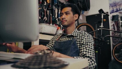 Asian man owner bike shop searching information gear by computer at cycle store. Young repairman set a selling price gear and accessory bike on website company. Small business and Owner shop concept