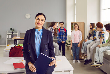 Portrait of female friendly cheerful teacher standing at school in classroom looking at the camera and smiling with a school children talking in background. Education, teaching concept.