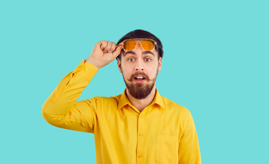 Stunned funny young bearded man isolated on blue studio background take off glasses shocked by deal or offer. Amazed male feel surprised with good seasonal promotion or discount.