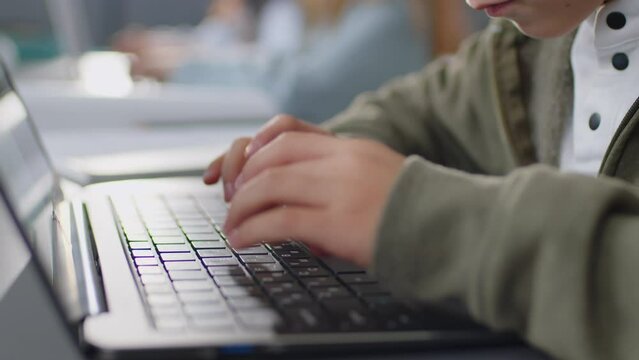 Focus on hands of elementary age boy sitting at desk in school classroom typing something on laptop keyboard