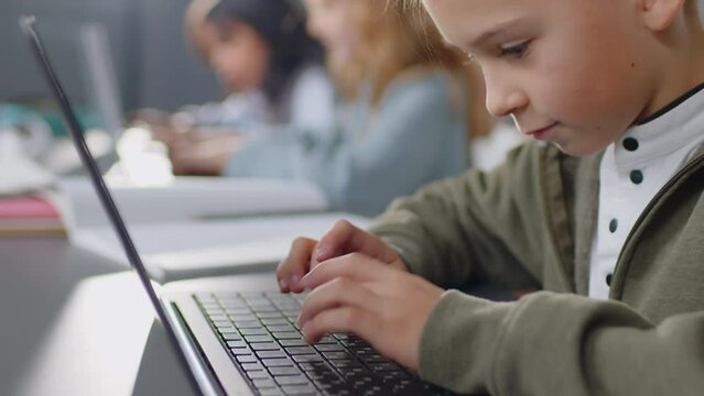 Tilt focus on Caucasian elementary age boy sitting at desk in classroom attentively typing something on laptop at daytime