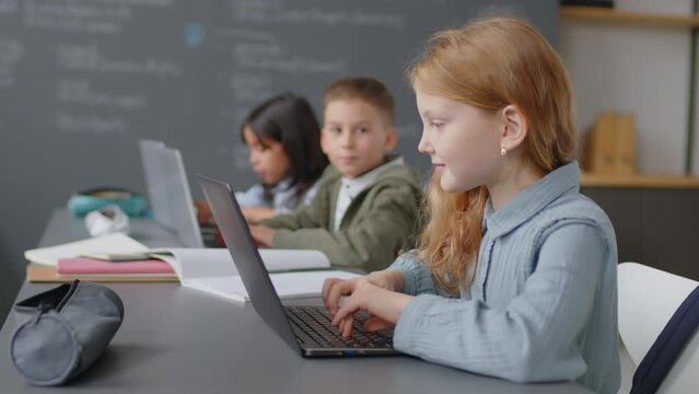 Medium shot of elementary age girl sitting at desk in school classroom typing something on laptop keyboard at daytime
