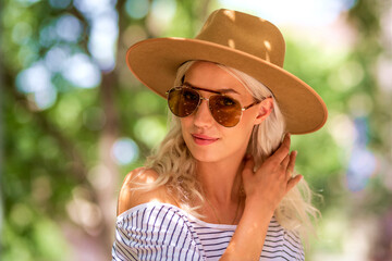 Stylish blond haired woman wearing hat and sunglasses while standing on the street