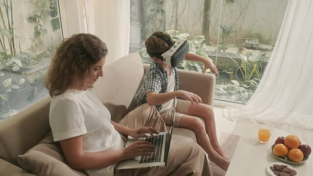 High angle view of son driving car on virtual glasses and mother working on laptop sitting on sofa in living room together