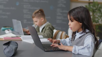 Selected focus on Hispanic schoolgirl sitting at desk in classroom typing something on laptop keyboard at daytime - Powered by Adobe