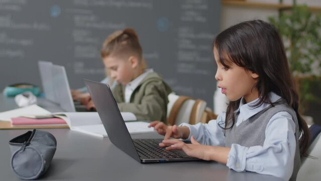 Selected focus on Hispanic schoolgirl sitting at desk in classroom typing something on laptop keyboard at daytime