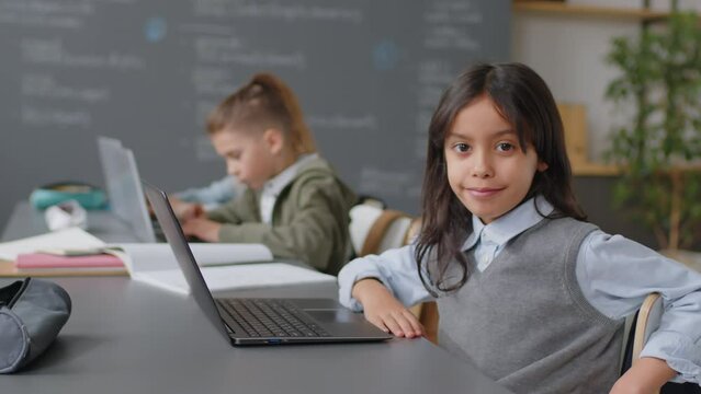 Portrait of Hispanic schoolgirl sitting at desk in classroom in front of laptop looking at camera at daytime