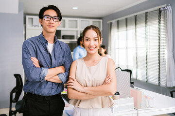 Cheerful young Asian businessman wearing eyeglasses and a businesswoman in casual smiling and looking at the camera while standing with her arms crossed. Happy businesswoman standing in the office.