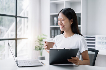 Asian businesswoman sits in a office working on laptop and enjoys a coffee.