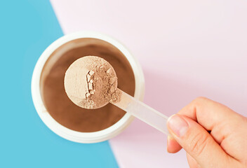 Chocolate protein powder in scoop and plastic jar on blue and pink background.