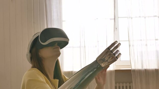 A woman in virtual reality glasses with a bionic arm prosthesis sits on a bed and moves her hands