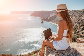 Digital nomad, woman in the hat, a business woman with a laptop sits on the rocks by the sea during sunset, makes a business transaction online from a distance. Freelance, remote work on vacation.