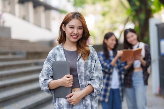 Happy young Asian woman student holding laptop on campus outdoor.