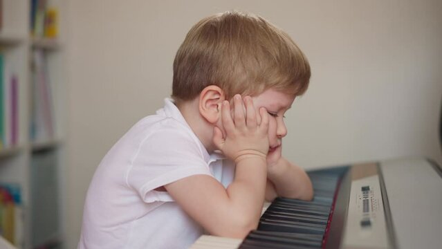 Little boy stops playing piano and props cheeks with hands putting elbows on keyboard failing to learn composition. Pensive toddler kid tired of difficult theme repeating