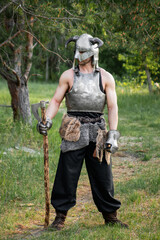 Portrait of a warrior in a horned helmet, a steel breastplate with a two-handed ax in his hands, posing against the backdrop of the forest. 