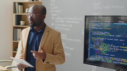 Medium close up of male African American teacher standing in front of blackboard in classroom giving lecture on programming at daytimeMedium close up of male African American teacher standing in front