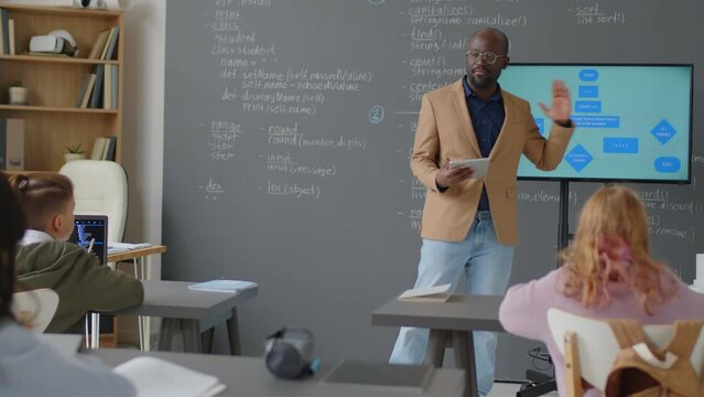 Full shot of male African American teacher standing in front of class holding tablet and explaining diagram on screen to elementary age students at daytime