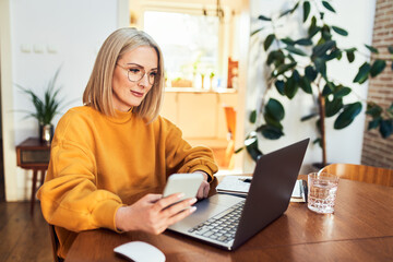 Portrait of adult woman using mobile phone while sitting in living room with laptop - obrazy, fototapety, plakaty