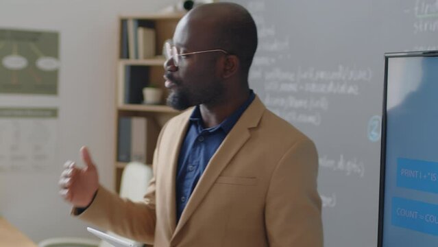 Tilt shot of male African American teacher standing in front of blackboard holding tablet and explaining diagram on screen to elementary age students at daytime