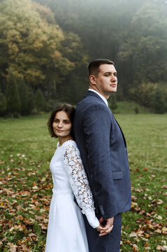 Wedding Walk In The Autumn Park. Newlyweds With A Difference In Height. Wedding Couple After The Ceremony. Couple Holding Hands Tightly.