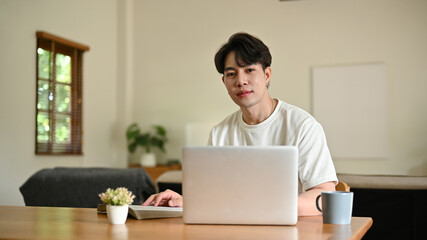 A handsome young Asian man sits at a table with his laptop in his minimalist living room.