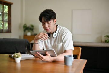 A focused young Asian male using his digital tablet at a table in his living room.